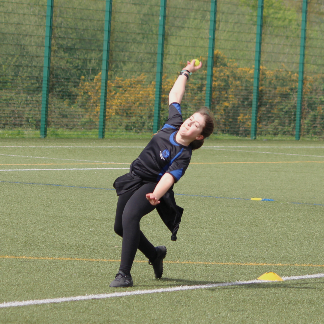 Pupil in PE kit bowls a cricket ball during a session on the school field, arm extended high in a bowling action.
