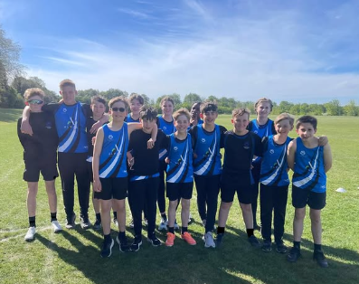 Internal alt text: A group of Year 7 and 8 pupils in blue and black Swanmore College athletics kit standing together on a grass athletics field during a sunny track and field event.