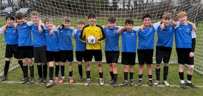 A large group of Year 7 boys stand shoulder to shoulder in front of a goal on an outdoor football pitch. They are wearing blue football shirts and black shorts, with a goalkeeper in a bright yellow jersey holding a football in the centre.