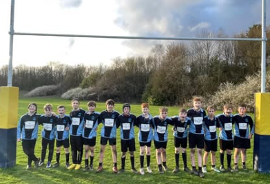 Year 7 boys rugby team standing in a line with arms around each other on a grass pitch beneath rugby posts after their match.