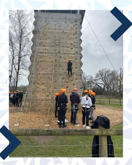 Image shows the full side of the  wooden climbing tower with one group of pupils on the ground in front of it, and one on the left side of the tower. there is a pupil abseiling down the wall.