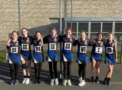 Seven Year 7 girls in Swanmore netball kit stand arm in arm on an outdoor court, smiling after a match with a school building and fencing behind them.