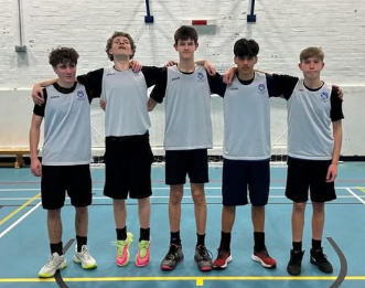 Five Year 11 boys stand in a line in a sports hall wearing white and black basketball tops with dark shorts. They have their arms around each other’s shoulders and are standing on a marked indoor court.