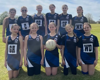The Year 7 girls netball team pose together outdoors on a field, wearing blue and black netball kits with position bibs, with one pupil holding a netball at the front.
