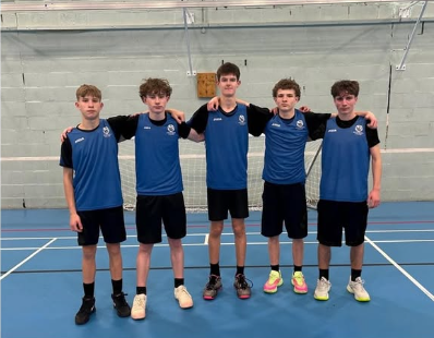 Image shows five U16 boys standing side by side in a sports hall, arms around each other’s shoulders. They are wearing matching blue and black Swanmore College basketball kit with black shorts and trainers. A netted goal and indoor court markings are visible behind them.