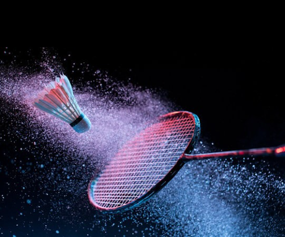 Image shows a shuttlecock about to be hit by a badminton raquet, against a dark background with a trail of powder or water coming from the raquet