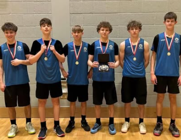 The Swanmore College Under 16 boys basketball team standing indoors wearing medals and holding a trophy after winning the District Plate Final.