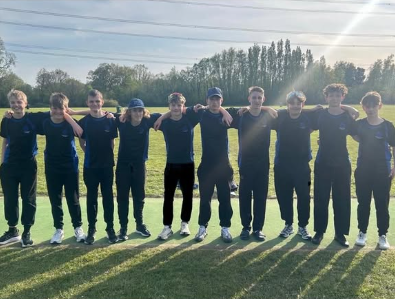 A group of boys from the Under 15 cricket team stand in a line on a grassy field with their arms around each other, wearing dark sports kit, with sunlight casting long shadows behind them.