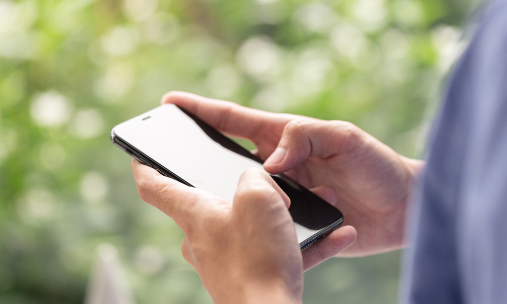 A person holding a smartphone with both hands, tapping the screen, with a soft green, out-of-focus background suggesting an outdoor setting.