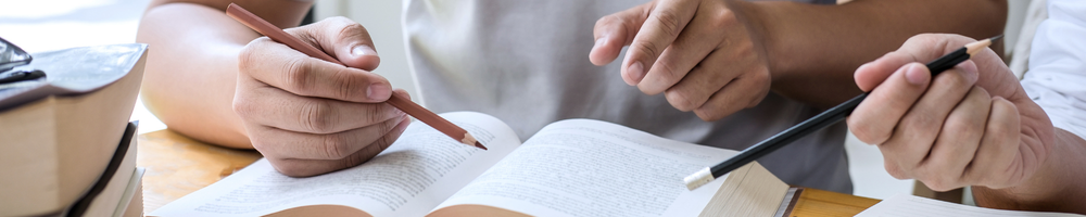 Image shows two pupils sitting at a desk pointing at an open textbook with pencils, appearing to discuss or revise the content together.