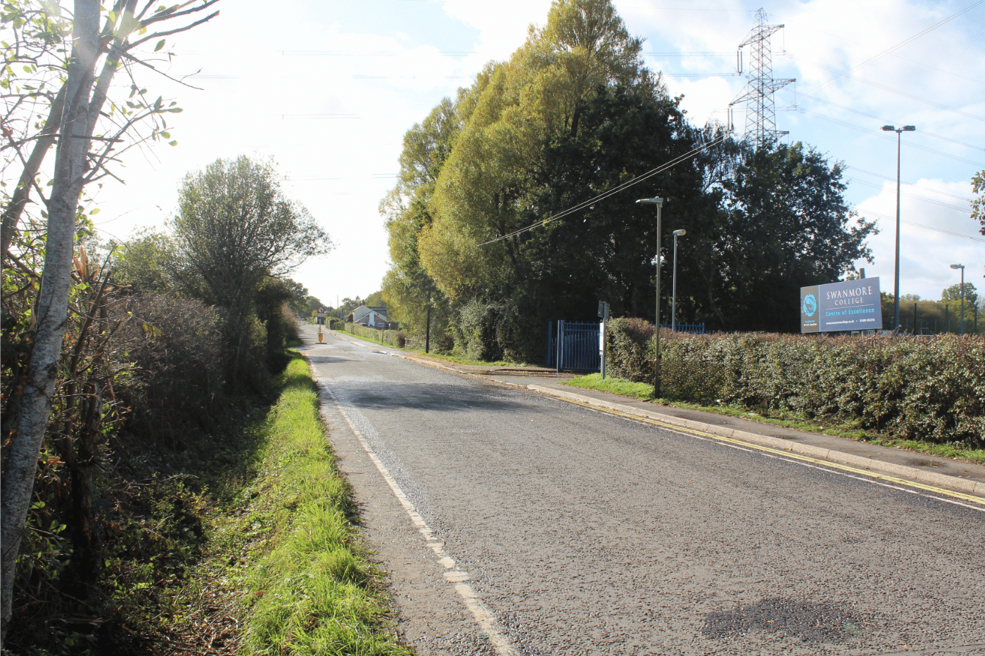 A view of New Road leading up to Swanmore College, showing the roadside entrance sign, trees, hedges, overhead wires, and an electricity pylon.