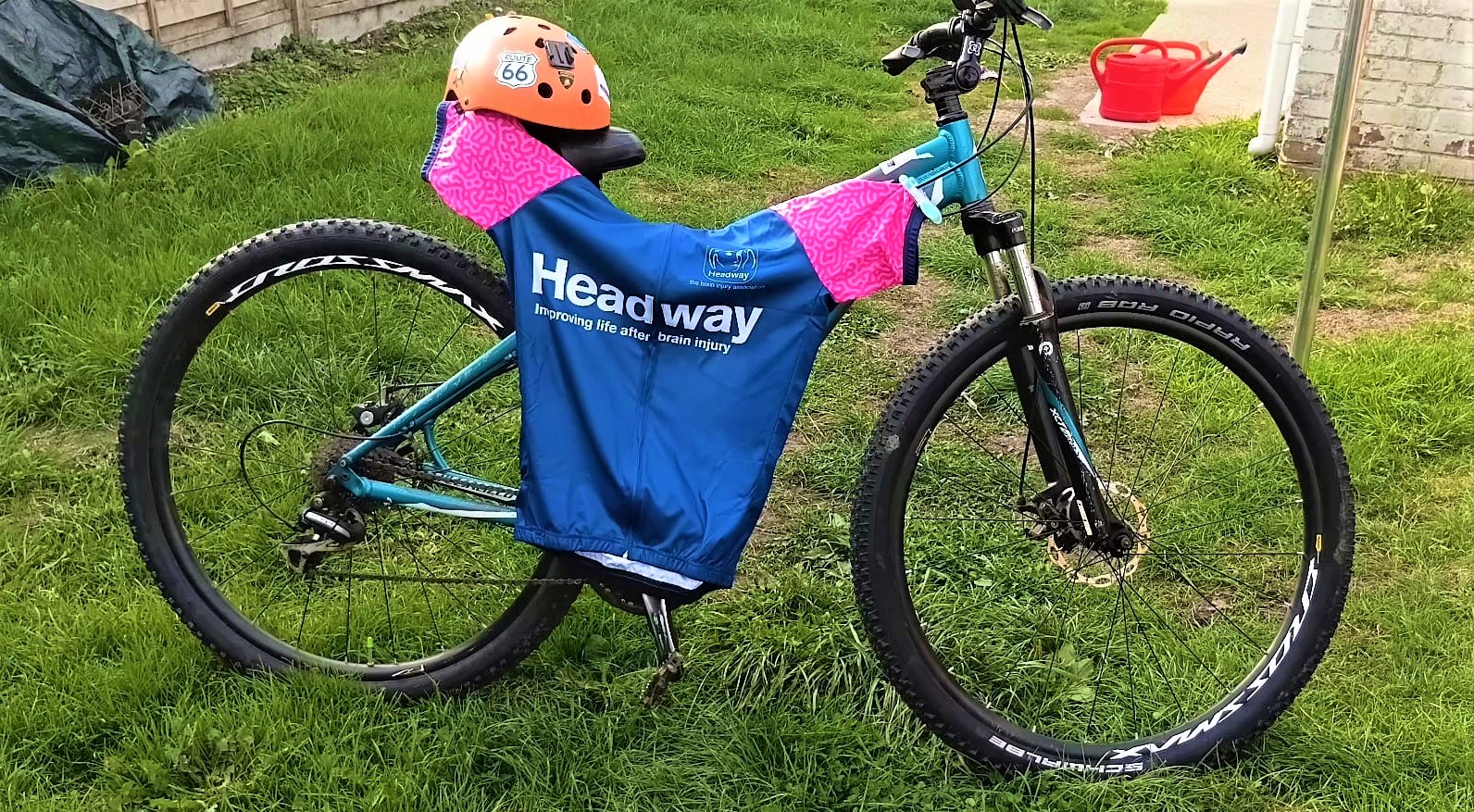 A blue mountain bike parked on a grassy lawn, draped with a blue and pink cycling jersey featuring the "Headway — Improving life after brain injury" logo and topped with an orange cycling helmet resting on the saddle.