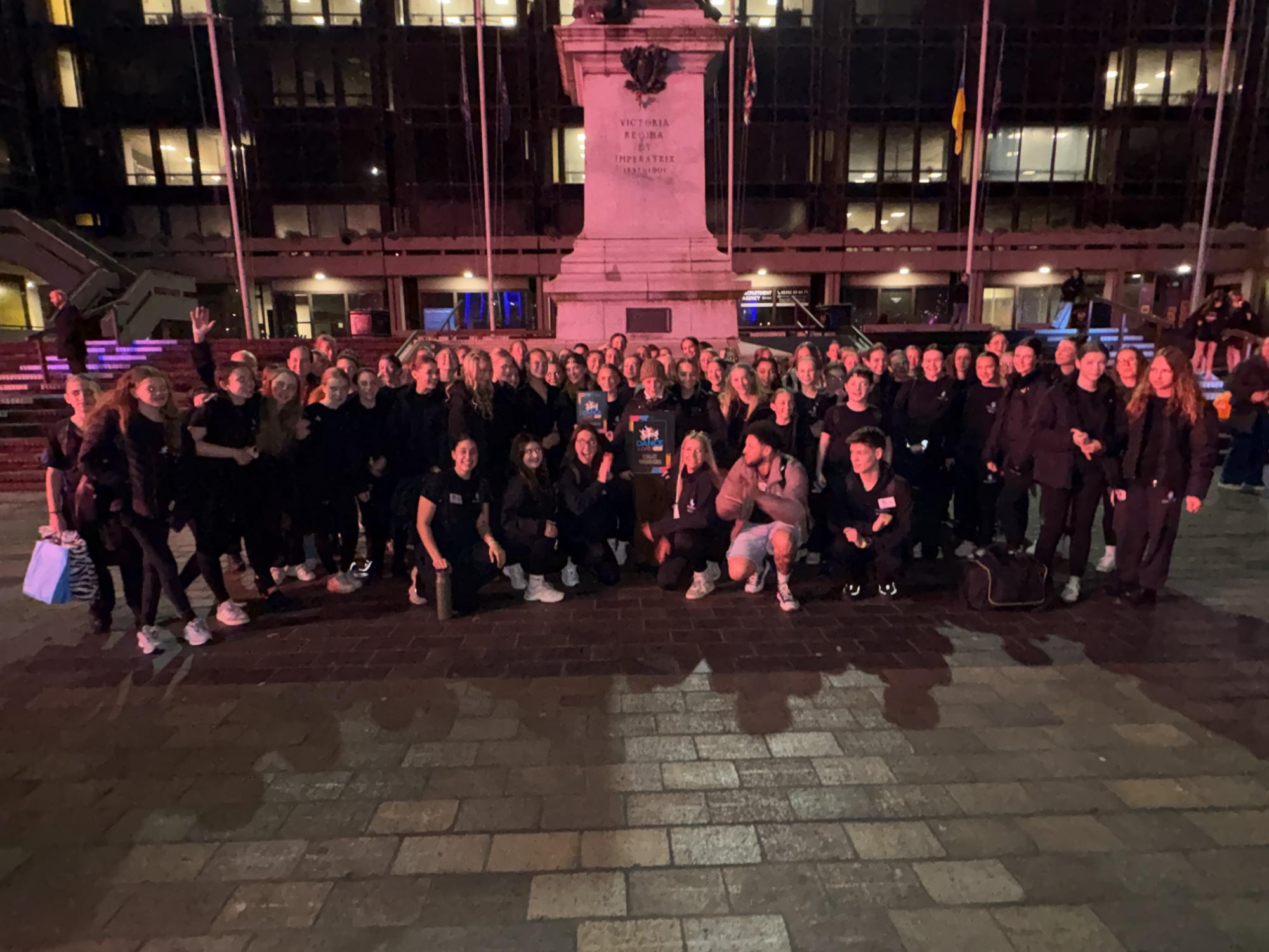 Image shows a large group of Swanmore College pupils and staff standing together outdoors at night in front of a city monument, wearing mostly black clothing and smiling after the Dance Live event.