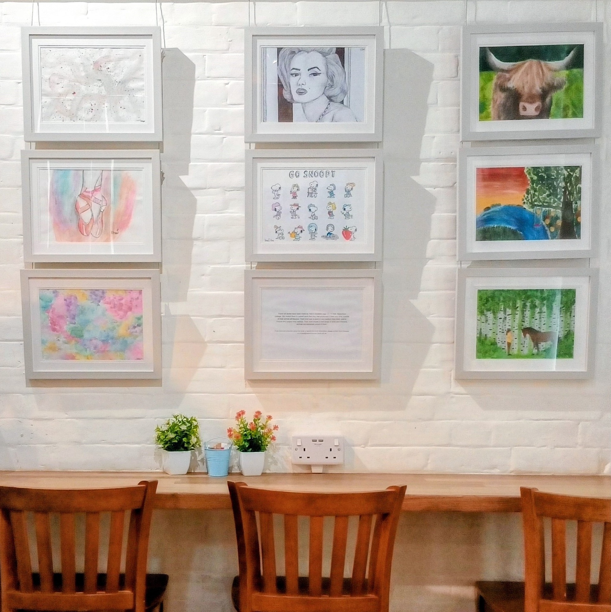Framed Year 8 pupil artwork displayed on a white brick wall above wooden café seating at St Barnaby’s Café in Swanmore.