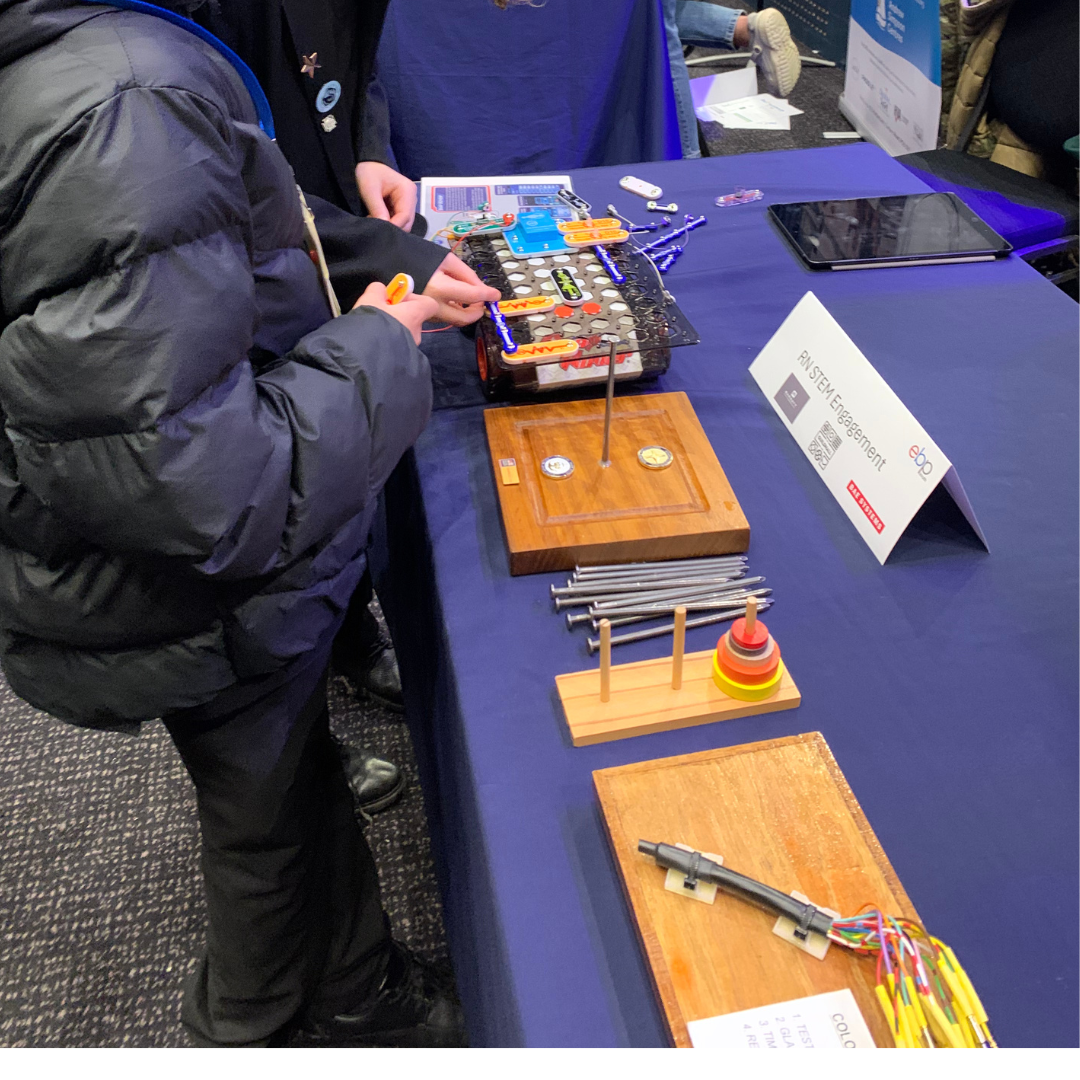 Pupils take part in a hands-on STEM activity at an engagement stand, using a construction kit with colourful components, circuits and tools on a table. Nearby are wooden learning resources, metal rods, stacking rings, cables and an iPad