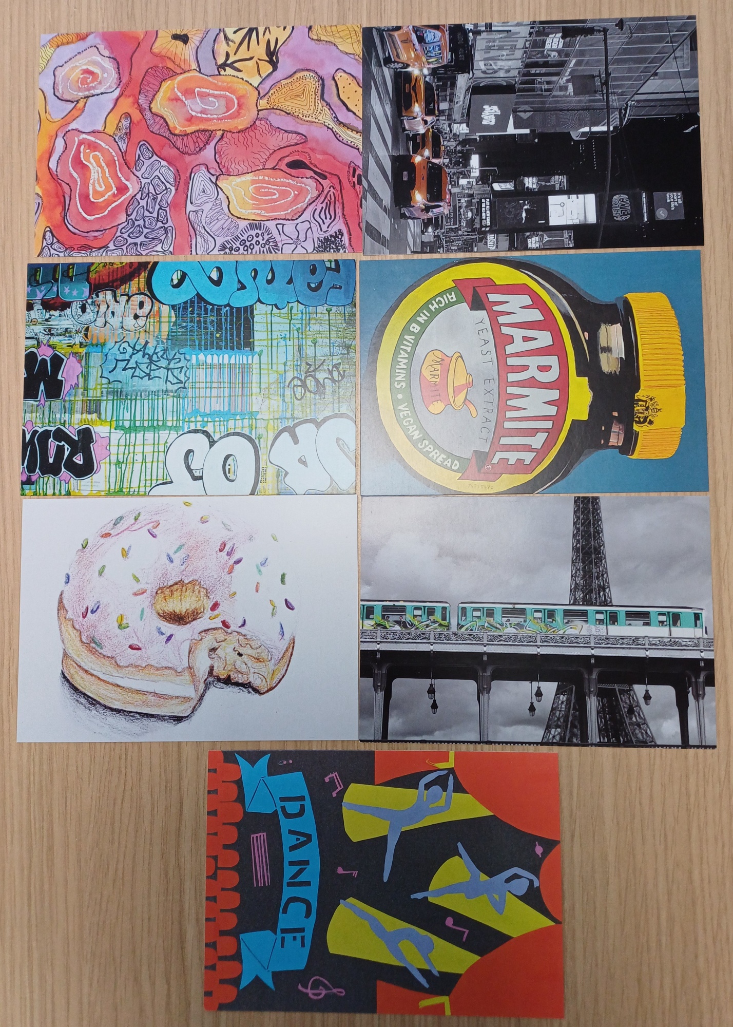 Seven postcard-sized art postcards arranged on a wooden table. Designs include abstract red and orange shapes, a black-and-white city street, colourful graffiti lettering, a Marmite jar illustration, a bitten iced doughnut with sprinkles, a train in front of the Eiffel Tower, and a bold dance-themed graphic with stylised figures.
