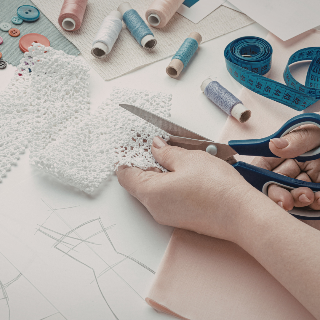 Image shows someone's hands cutting a strip of lace with scissors. Their work station is surrounded by reels of thread, measuring tape and other paraphanalia