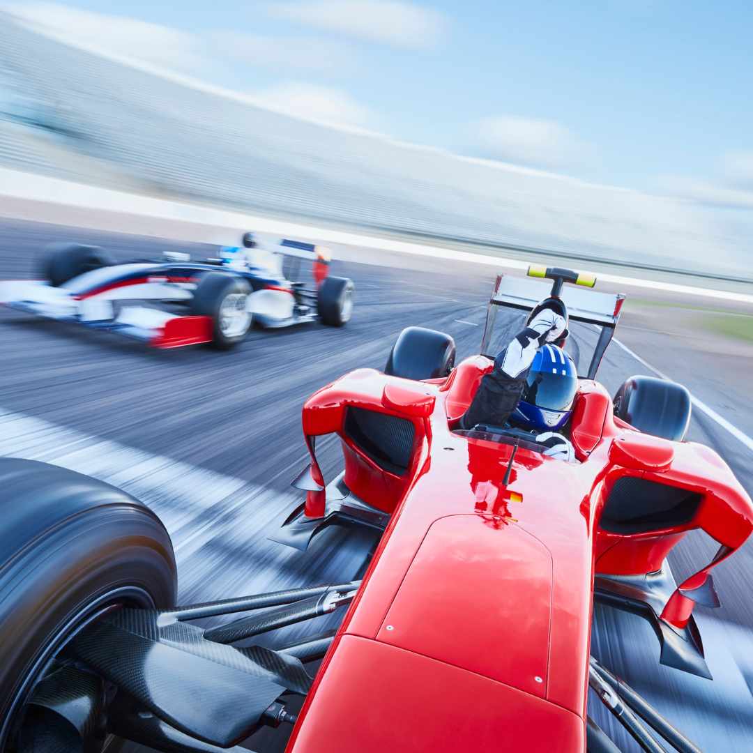 Image shows a picture of a victorious racing car driver waving his fist in the air, whilst still driving, with another car behind and the stands a motion blur