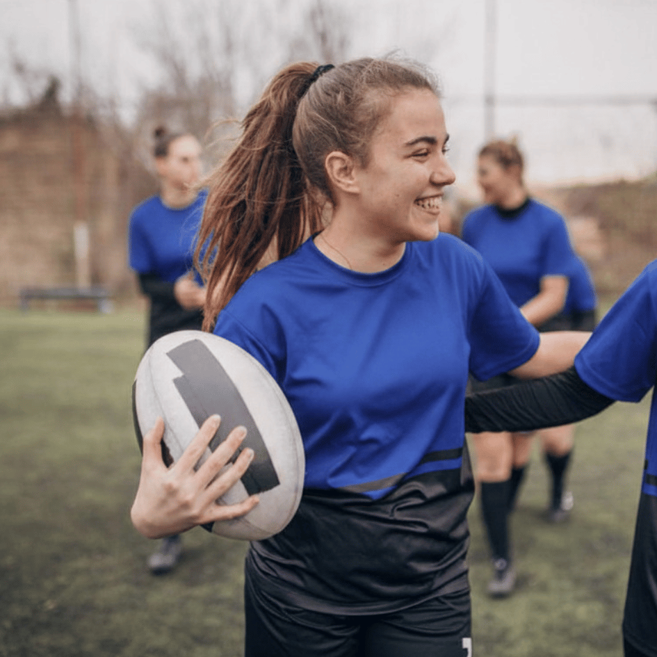 Image shows a teenage girl holding a rugby ball with one arm, her other arm is around a friend who is just out of the picture, and she is smiling at her.