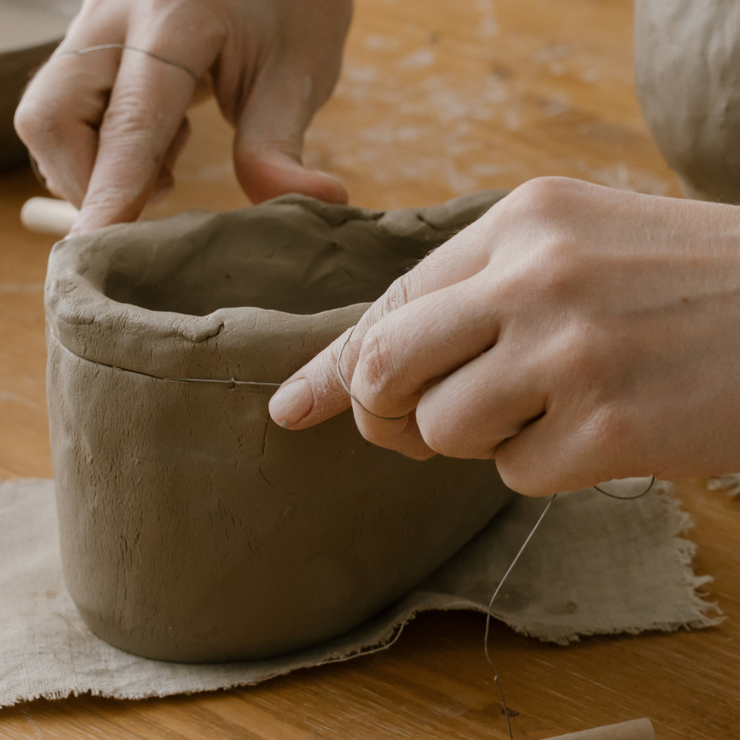 Image shows a pair of hands using a cheese wire to remove the uneven top of an unfired clay pot