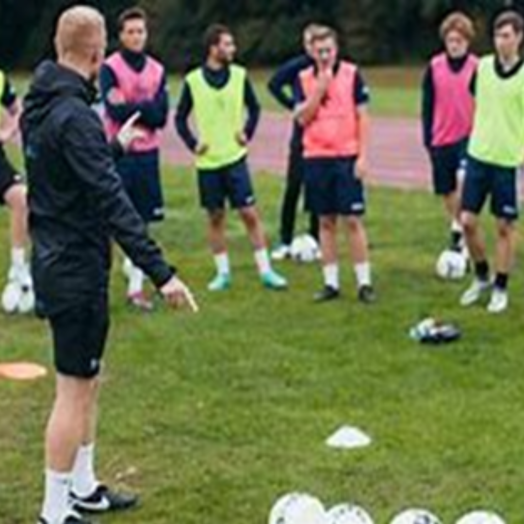 Image shows a sports coach surrounded by people with a pile of footballs at his feet.