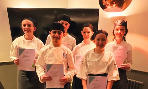 Six pupils in white chef uniforms and hats stand together holding certificates after graduating from the Eastleigh College Junior Chef course.