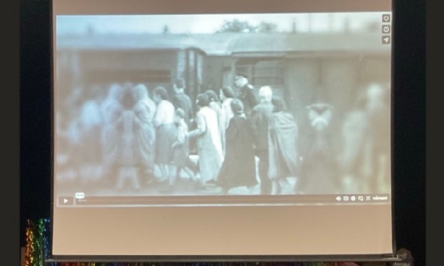 Image shows a projected black-and-white historical film clip of a group of people wearing coats, gathered closely together beside railway carriages.