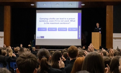 Students seated in a school hall raise their hands during an assembly as a police officer presents a question on prison sentences for carrying a knife