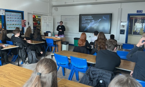 Adult presenter points to a classroom screen showing an aerial image and diagrams of the Havant Thicket Reservoir, with pupils seated facing forward.