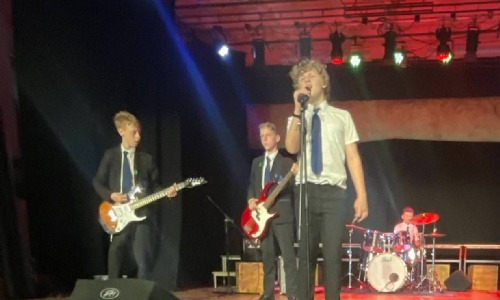Image shows KS3 pupils performing music on stage - there are 4 pupils, 1 on drums in the background, 2 on electric guitar and 1 singing at the front.