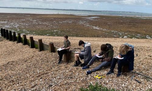 Pupils sit on a pebble beach at Hill Head collecting fieldwork data and recording observations in notebooks.