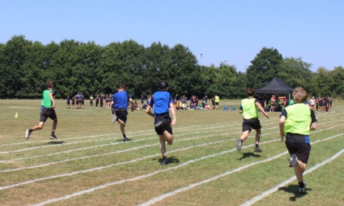 Pupils sprint along the running track during a relay race at Sports Day, with spectators and staff gathered beside the field.