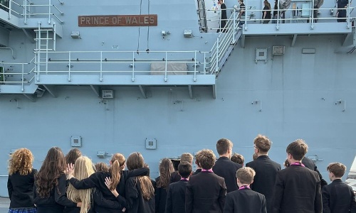 Pupils in uniform look up at a ‘PRINCE OF WALES’ sign mounted on the side of a grey naval ship.